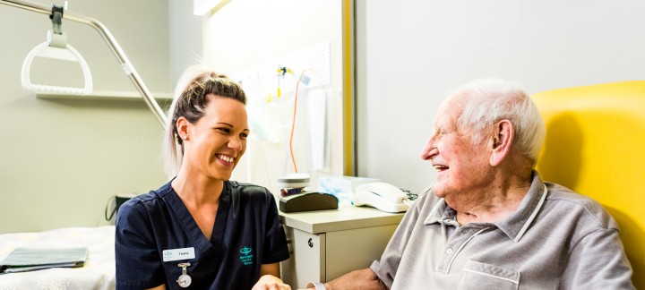 An elderly man sits in a yellow chair at a table with a bowl of fruit, holding the hand of a young female Mercy Health worker. The man and woman are smiling at each other.