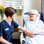 A Mercy Health female worker with short brown hair sits to the left of an elderly resident who holds her hand. They smile at each other.