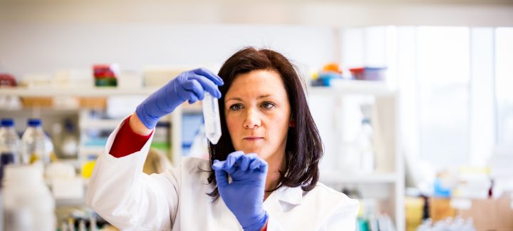 A medium close up of a woman in a lab wearing blue gloves and white lab coat holding a test tube.