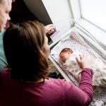 A man and a woman Look down at a new born baby wrapped up and asleep in a hospital crib, the woman rests her hand on the baby.