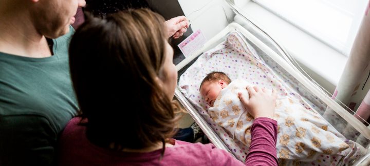 A man and a woman Look down at a new born baby wrapped up and asleep in a hospital crib, the woman rests her hand on the baby.