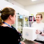 A woman holds a styrofoam box while smiling at another woman in a lab room