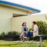 Two women sit on a metal park bench in a small garden at Mercy Health talking, one woman is smiling.