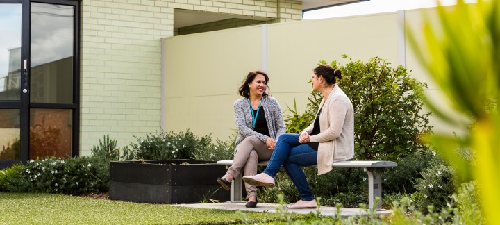 Two women sit on a metal park bench in a small garden at Mercy Health talking, one woman is smiling.