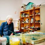 An elderly man with curly hair sits in an art room and looks at the camera. He is surrounded by canvases and paints.