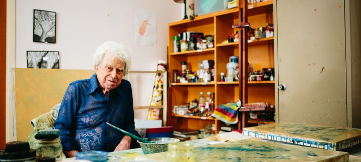 An elderly man with curly hair sits in an art room and looks at the camera. He is surrounded by canvases and paints.
